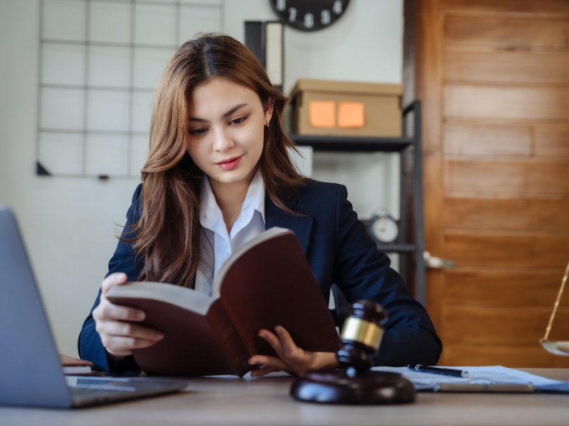 A young woman studying from a book, with a gavel in front of her and a laptop.
