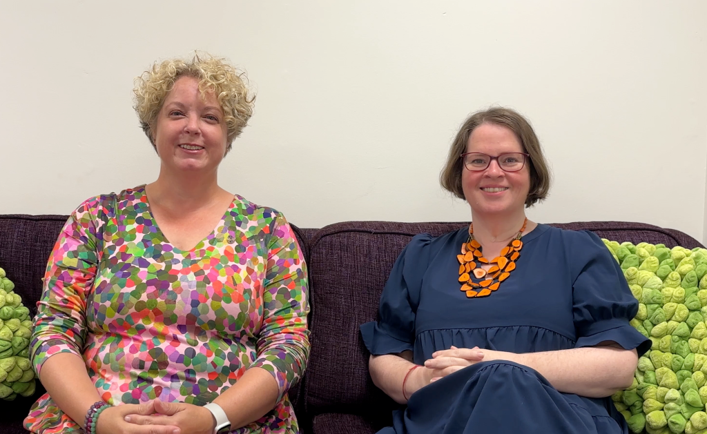 Two women sit on a purple sos with green pillows in front of a blank wall