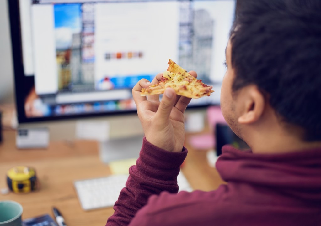 Businessman, eating and lunch with computer screen in office with rear view and pizza break for working on deadline. Programmer, employee and back with fast food, coding display or hungry at desk