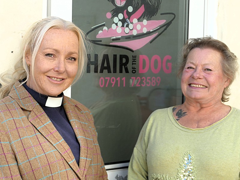 Two women stand in front of a dog groomers door, one women has a clerical collar