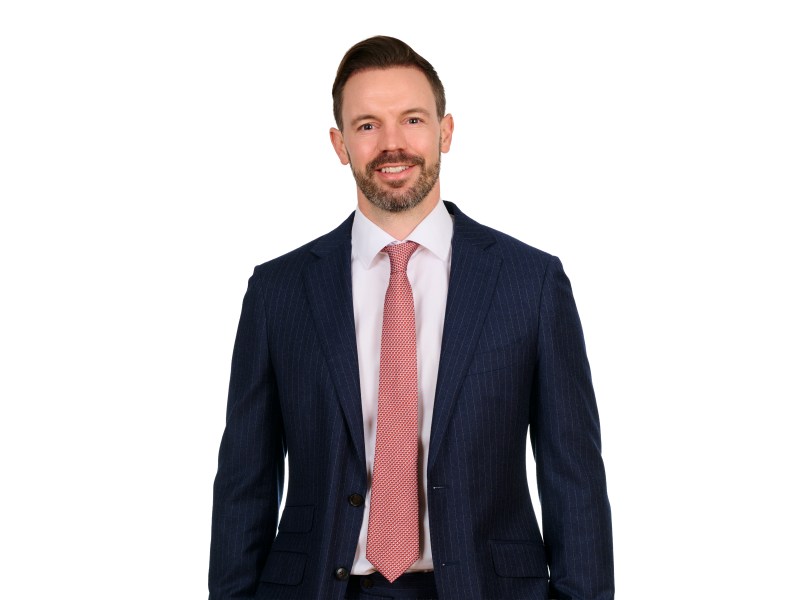 A man stands in front of a white background in a dark suit and red tie