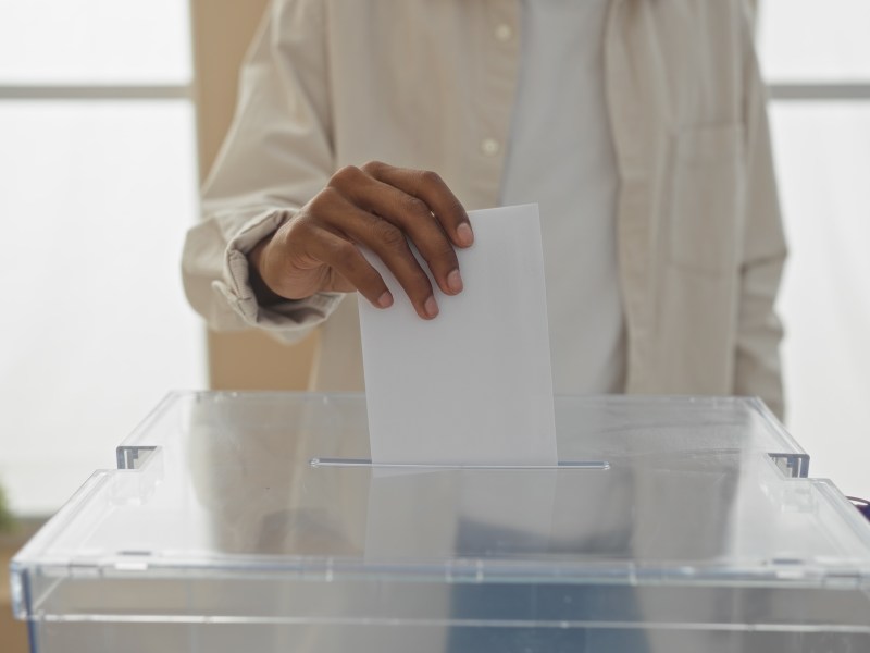A man casting a vote by placing a ballot into a transparent box during an election held indoors.